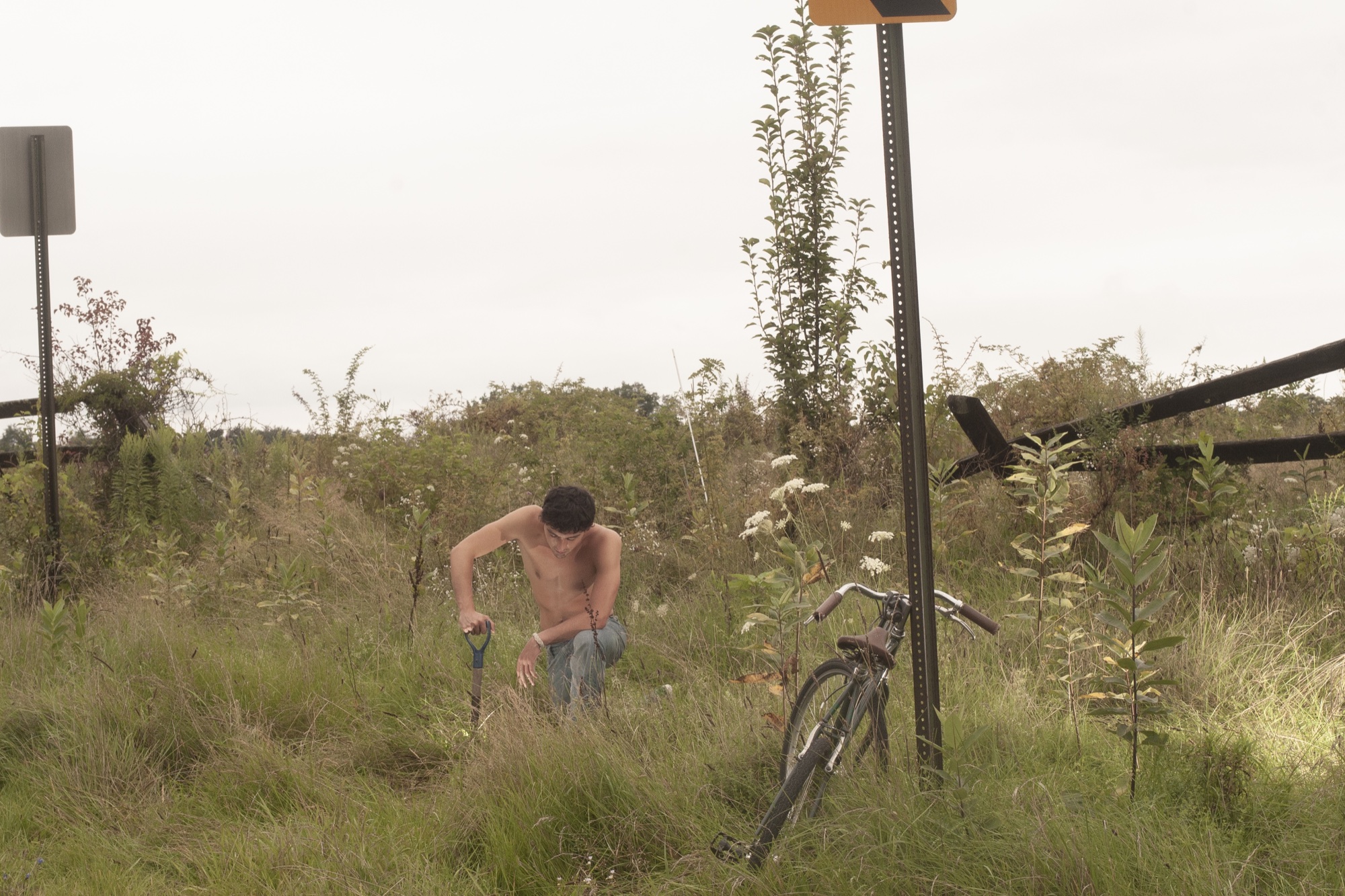 Figure crouching in tall grass beside a bicycle and roadside signposts