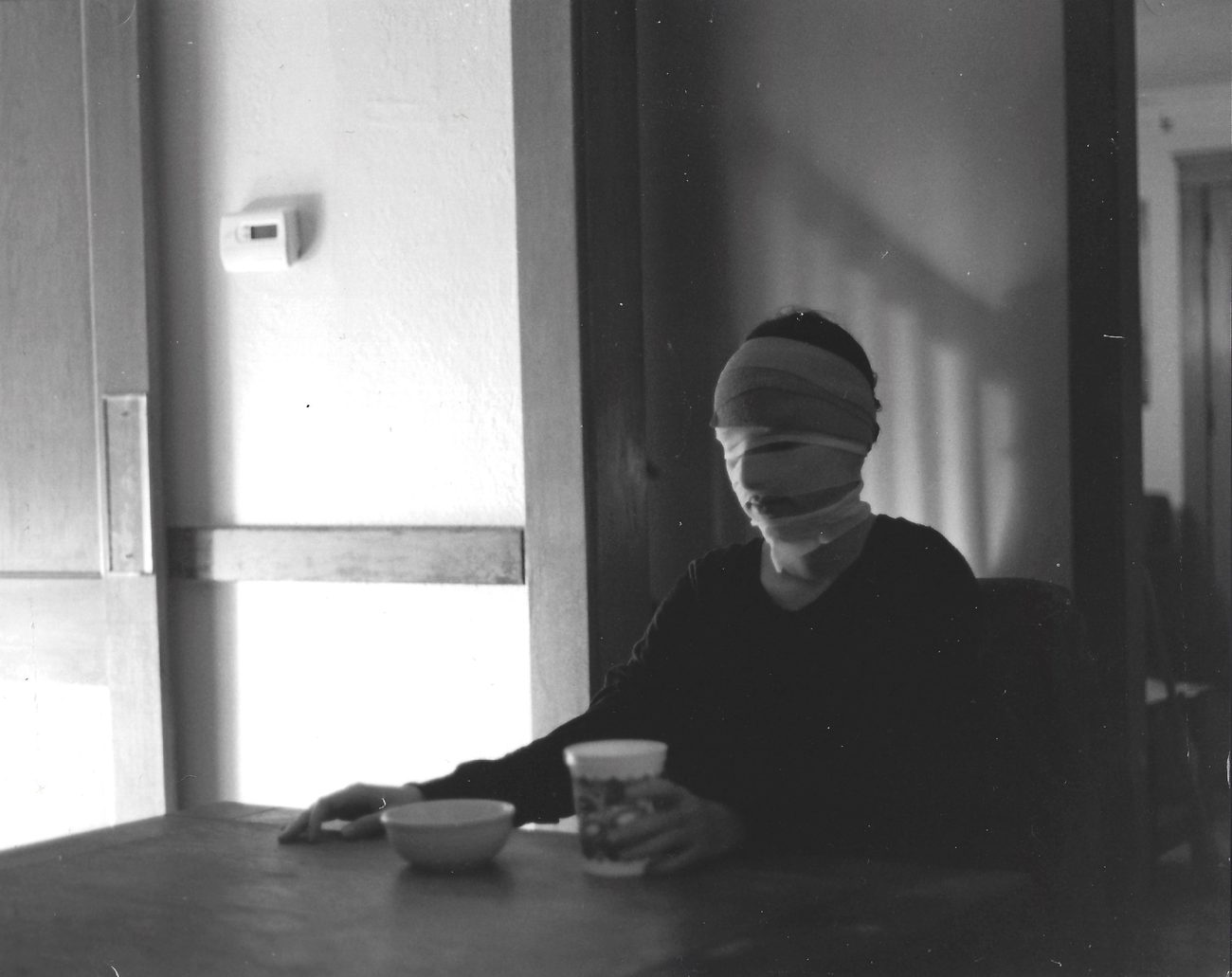 Black and white interior: figure seated in a darkened room beside a doorway