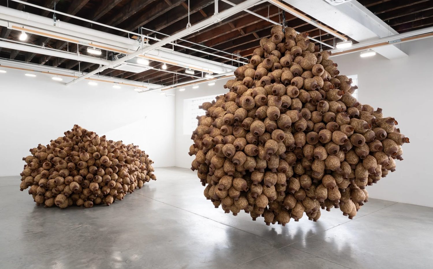 Gallery installation: a large suspended cluster of ceramic vessel forms, with a second mound resting on the floor